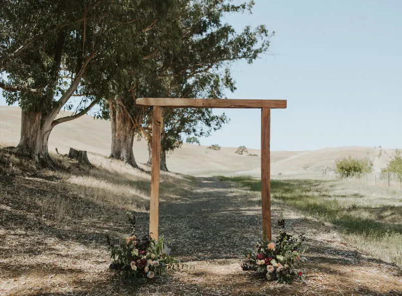 A wooden ceremony arch standing in a golden field beneath a spreading oak at Glenn Ranch