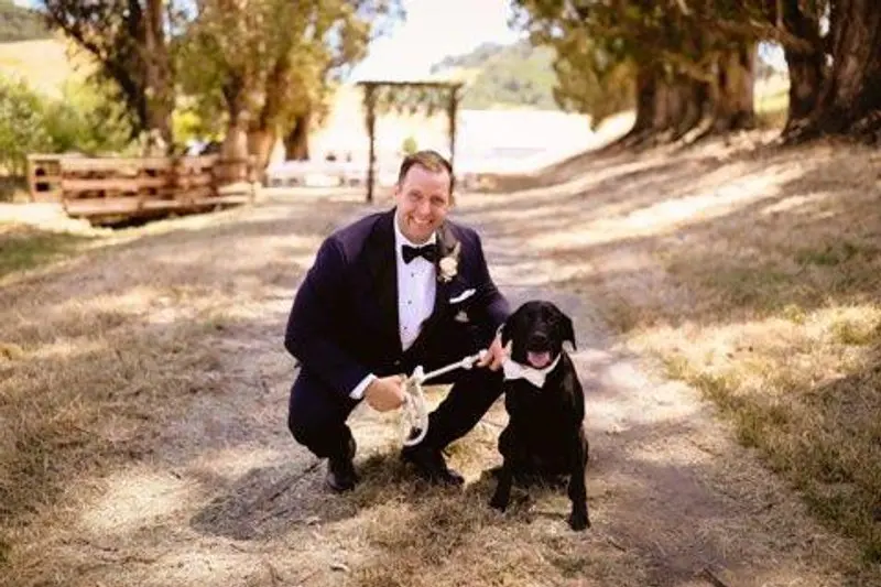 A groom kneeling with the family dog on the oak-lined drive