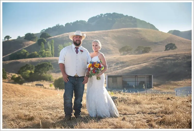 A couple standing in the rolling golden hills with a red barn behind them at Glenn Ranch